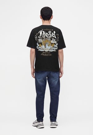 Hombre de espaldas con camiseta negra Diesel con un gran gráfico ornamentado y texto, vaqueros azules y zapatillas blancas sobre fondo liso.