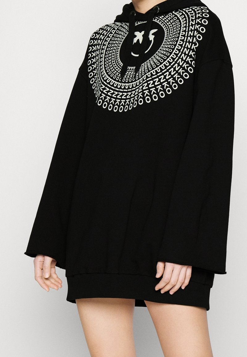 Personne portant un sweat à capuche noir oversize avec un motif de texte circulaire blanc et un design de visage souriant autour de l'encolure.