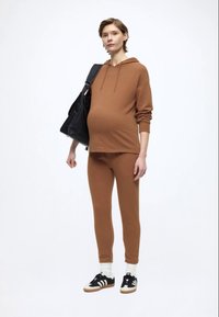 Ruda džemperis su gobtuvu ir atitinkamos tamprės iš minkštos medžiagos; modelis neša juodą rankinę; baltos kojinės ir juodi sportbačiai su trimis juostelėmis.