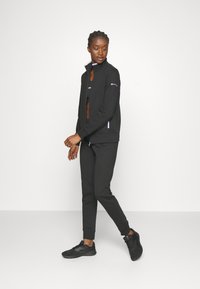 Chaqueta deportiva negra con cremallera y alto cuello, de manga larga, y acentos morados contrastantes. Combinada con pantalones jogging negros a juego y zapatillas deportivas.