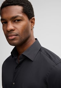 Primo piano di un uomo che indossa una camicia nera con colletto aperto e un piccolo orecchino ad anello d'oro nell'orecchio sinistro.