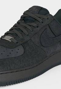 Χαμηλοτάκουνο μαύρο αθλητικό παπούτσι Nike Air Force 1 με υφασμάτινη υφή, μαύρα κορδόνια, διάτρητο μπροστινό μέρος και μεταλλικό ταμπελάκι κορδονιών σε λευκό φόντο.