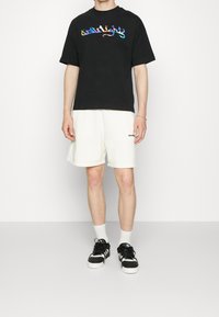 Junge Person in einem schwarzen T-Shirt mit buntem Text, weißen Shorts, weißen Socken und schwarz-weißen Turnschuhen, die vor einem weißen Hintergrund steht.