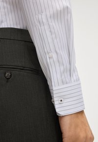 Polsino di camicia bianca a righe con dettagli dei bottoni, abbinato a pantaloni grigi scuri con texture e una tasca visibile. Stile formale classico.