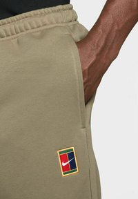 Beige sporta bikses ar elastīgu jostasvieta, kurām ir neliela Nike logo apdare krāsainā kvadrāta dizainā kreisajā augšstilba kabatā.