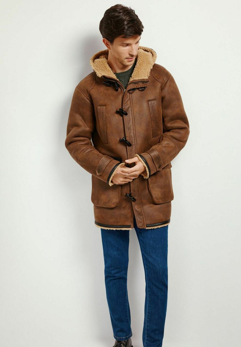 Brauner Ledermantel mit einer Kunstfellkapuze, Knopfverschlüssen, zwei vorderen Taschen und strukturierten Ärmeln; kombiniert mit blauen Jeans.