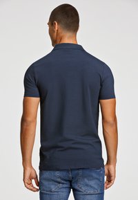 Camisa polo azul marino de tela suave, con cuello y mangas cortas, combinada con jeans de mezclilla, mostrando una vista trasera sencilla.