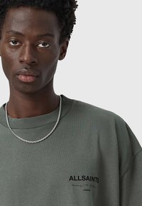 Sudadera verde oliva de corte oversized con un cuello redondo acanalado, que presenta un pequeño logo negro en el pecho y una cadena de plata.