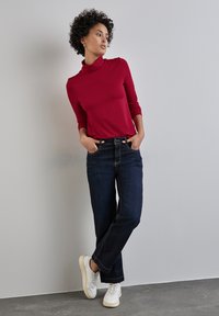 Röd polotröja, mörkblå denimjeans och vita sneakers. Slät textur med en figursydd design. Avslappnad outfit mot en neutral bakgrund.