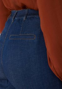Jeans in denim blu con vita alta, dotati di una tasca cucita sul retro e una texture liscia, abbinati a un top color ruggine.