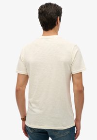 Camiseta de manga corta de color crema con una textura suave, corte relajado, cuello redondo y un sutil patrón jaspeado. Se llevan vaqueros.