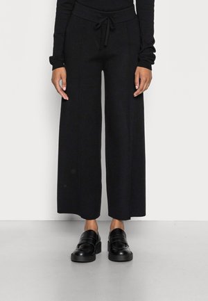 Persona que lleva pantalones negros de pierna ancha con cintura de cordón y mocasines negros sin cordones, de pie sobre un suelo gris frente a una pared blanca.
