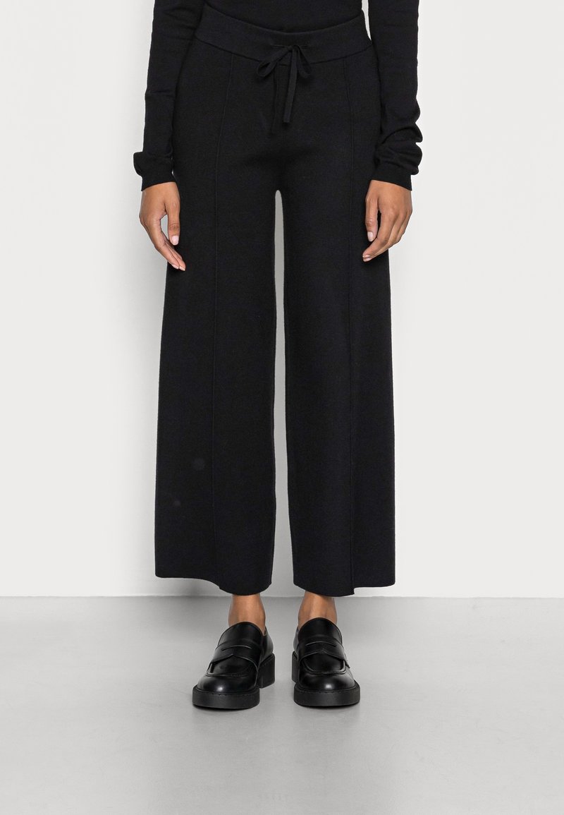 Personne portant un pantalon noir à jambes larges avec une taille à cordon et des mocassins noirs sans lacets, debout sur un sol gris contre un mur blanc.