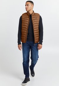 Braune gesteppte Weste mit Stehkragen, kombiniert mit einem marineblauen Pullover und blauen Jeans. Die Weste verfügt über einen Reißverschluss und ein Logo-Detail.