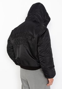 Zwarte bomberjack met capuchon en een gequilt ontwerp, voorzien van geborduurde "SCHOTT NYC" tekst op de achterkant, geribde cuffs en onderkant.