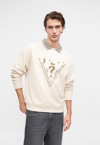 Ungdommelig mann iført en beige hettegenser med Guess-logo og grå krage, kombinert med mørkegrå jeans, stående mot en enkel hvit bakgrunn.