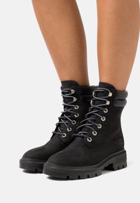 Botas de ante negro con cordones, cuello acolchado, ojales metálicos y suela de goma robusta con acentos texturizados.