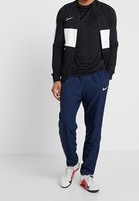 Nike-träningsdräkt bestående av en svart jacka med vita accenter, svart t-shirt och marinblå byxor, tillsammans med vita och röda träningsskor.