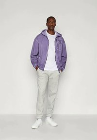 Casaco com fecho éclair roxo e capuz, com bolsos frontais, sobre uma t-shirt branca, combinado com calças de treino cinzentas claras e ténis brancos. Design simples.