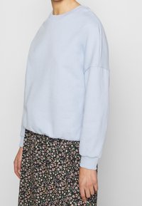 Felpa azzurra chiara in materiale morbido con polsini e scollatura a costine, abbinata a una gonna nera con motivo floreale caratterizzata da piccoli fiori multicolori.
