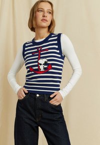 Maglione senza maniche a righe blu navy e bianche con un disegno di unancora e un cane cartone animato, abbinato a jeans in denim scuro. Texture liscia.