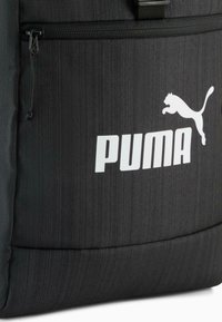 Sac à dos noir en tissu texturé avec un grand logo Puma blanc et une poche zippée à l'avant. Couture d'accentuation visible.
