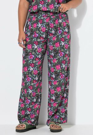 Person iført vidde bukser med et farverigt blomsterprint i pink, lilla og grøn, kombineret med grønne platformssandaler og ringe på fingrene.