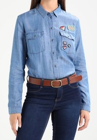 Donna che indossa una camicia di jeans azzurro chiaro con toppe e jeans blu scuro, accessoriate con una cintura di pelle marrone con fibbia in ottone.