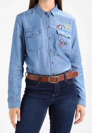 Kvinde iført en lyseblå denimskjorte med lapper og mørkeblå jeans, accessoriseret med et brunt læderbælte med messingspænde.