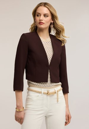 Blondynka w ciemnym krótkim blezerze, beżowym wzorzystym swetrze, białych spodniach z złotym łańcuszkowym paskiem, złotych kolczykach i bransoletce, patrzy w lewo.