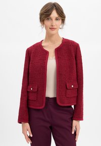 Donna che indossa un giubbotto rosso textured con tasche frontali e pantaloni borgogna, in piedi davanti a uno sfondo bianco semplice.