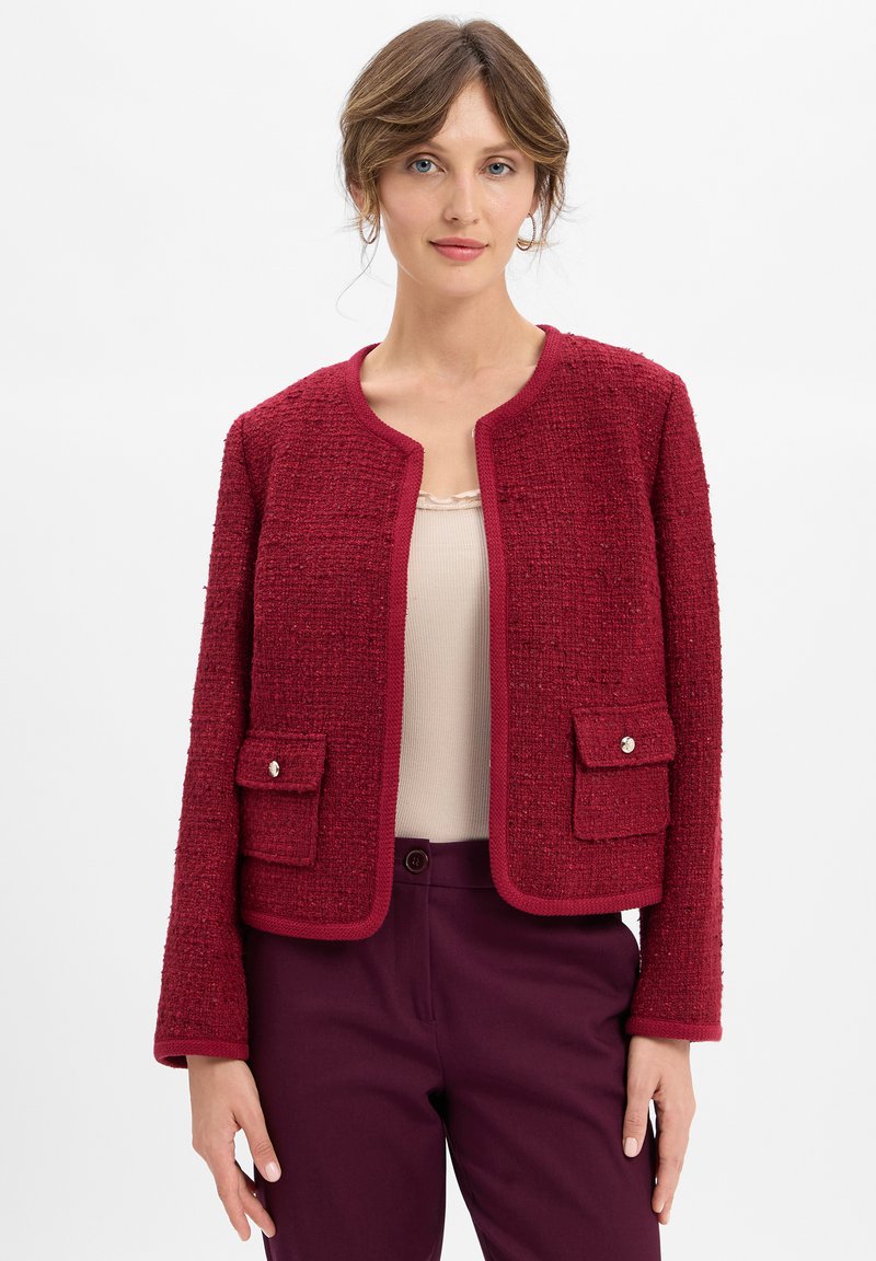 Donna che indossa un giubbotto rosso textured con tasche frontali e pantaloni borgogna, in piedi davanti a uno sfondo bianco semplice.