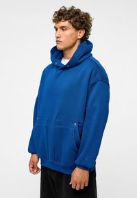 Blauwe oversized hoodie gemaakt van een zachte stof, met een grote voorkantzak, een capuchon met trekkoord en ribbenmanchetten. Soepele textuur met minimale details.
