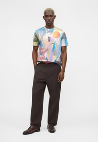 Mann med kort blond hår iført en fargerik abstrakt mønstret kortermet skjorte, mørkebrune bukser og brune pyntesko, står med hendene i lommene.