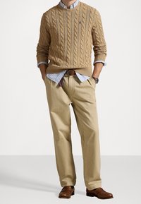 Maglione beige a trecce con polsini a coste, camicia button-up a righe blu chiaro sotto, pantaloni khaki pieghettati e scarpe eleganti marroni.