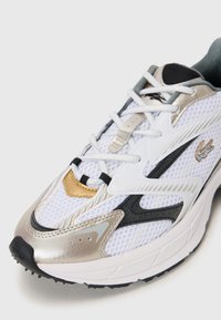 White athletic shoe with mesh upper, silver and black accents, pronounced sole, and gold detailing on the toe. Laces are white.
