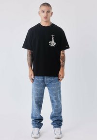 Zwarte katoenen t-shirt met een wit grafisch ontwerp op de borst, gecombineerd met blauwe denim jeans met een logo-print over de hele stof, en witte sneakers.