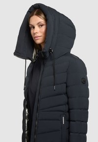 Donkerblauwe pufferjas met een grote capuchon, doorgestikte textuur en ritssluiting. Heeft zwarte koordjes en een logo op de mouw.