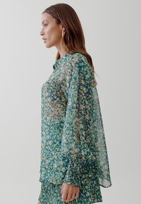 Sheer green blouse with floral pattern, long balloon sleeves, buttoned collar, and elasticized cuffs. Lightweight and flowing fabric.
