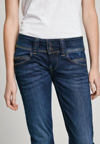 Mörkblå denimjeans med en klassisk design, två knappar och kontrasterande sömmar. Fickor är synliga på båda sidor.