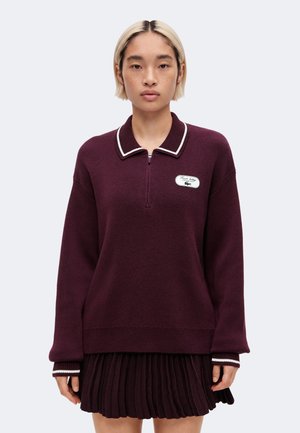 Jeune personne aux cheveux blonds courts portant un pull à manches longues violet foncé avec un bord blanc et une jupe plissée, debout face à l'avant.