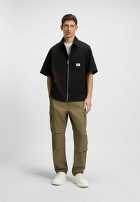Camicetta nera a maniche corte con zip e due tasche frontali, abbinata a pantaloni cargo verde oliva con molte tasche, indossata con sneaker bianche.