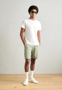 Witte t-shirt, lichtgroene linnen shorts, witte sneakers met grijze accenten en witte crew-sokken. Model staat met handen in de zakken.