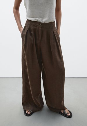 Personne portant un pantalon en lin marron ample, un haut blanc rentré, les mains dans les poches, et des sandales noires sur un sol gris clair.