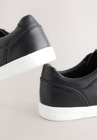 Paire de baskets en cuir noir avec semelles en caoutchouc blanches vues du talon sur un fond clair.