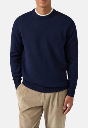 Uomo con maglione blu navy girocollo indossato sopra una camicia bianca, abbinato a pantaloni beige in velluto a coste, mani comodamente nelle tasche.