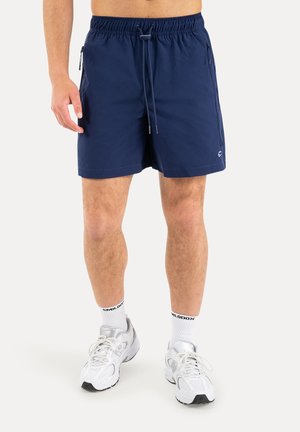 Man draagt marineblauwe shorts met trekkoord, witte sokken met zwarte tekst en witte sportschoenen, staand tegen een effen achtergrond.