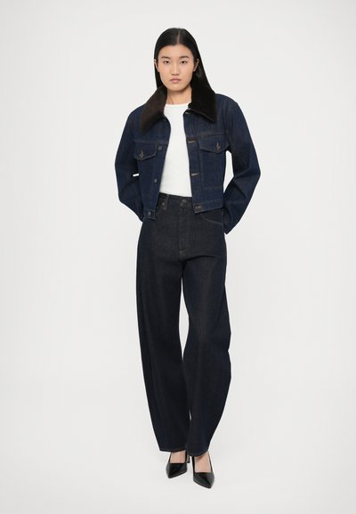 Tmavomodrá rifľová bunda s hnedým golierom z umelého kožúšku, v kombinácii s tmavomodrými rifľami s vysokým pásom a čiernymi lodičkami s ostrou špičkou.