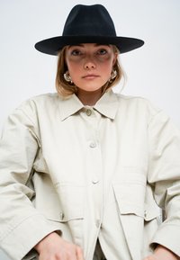 Helle beigefarbene Jacke mit Kragen, zwei Fronttaschen und silbernen Knöpfen. Getragen mit einem breitkrempigen schwarzen Filzhut und Creolen.