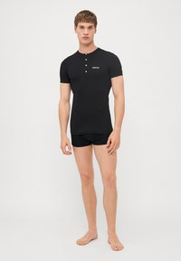 Schwarzes Kurzarmhemd mit Knopfdetails und Logo auf der Brust, kombiniert mit schwarzen Unterhosen. Glattes Material, enganliegendes Design, minimale Akzente.